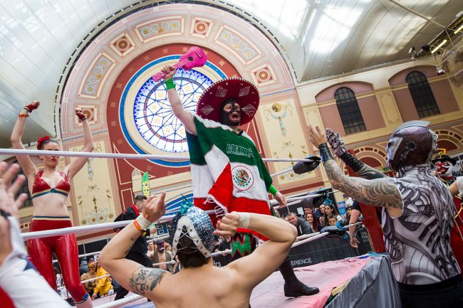 Person in sombrero and Mexican flag poncho wielding a pink ukelele at a crowd of Mexican wresters.
