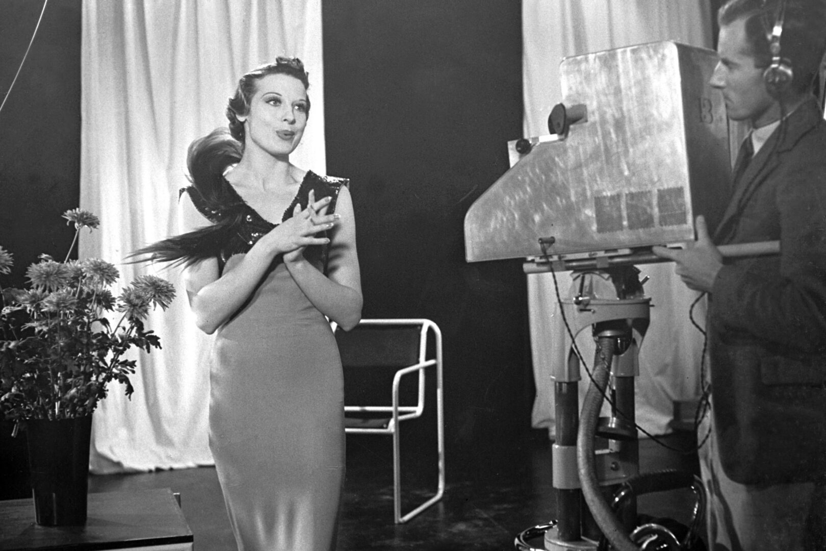 Glamourous vintage woman in dress with feather collar being filmed by man with a large camera.