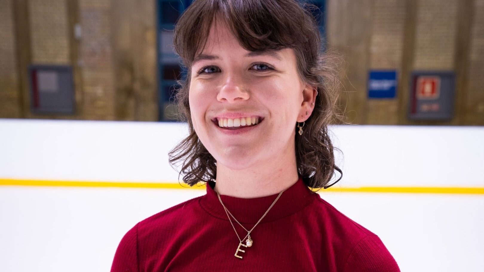Woman with brown hair in a red polo neck stands on ice rink smiling