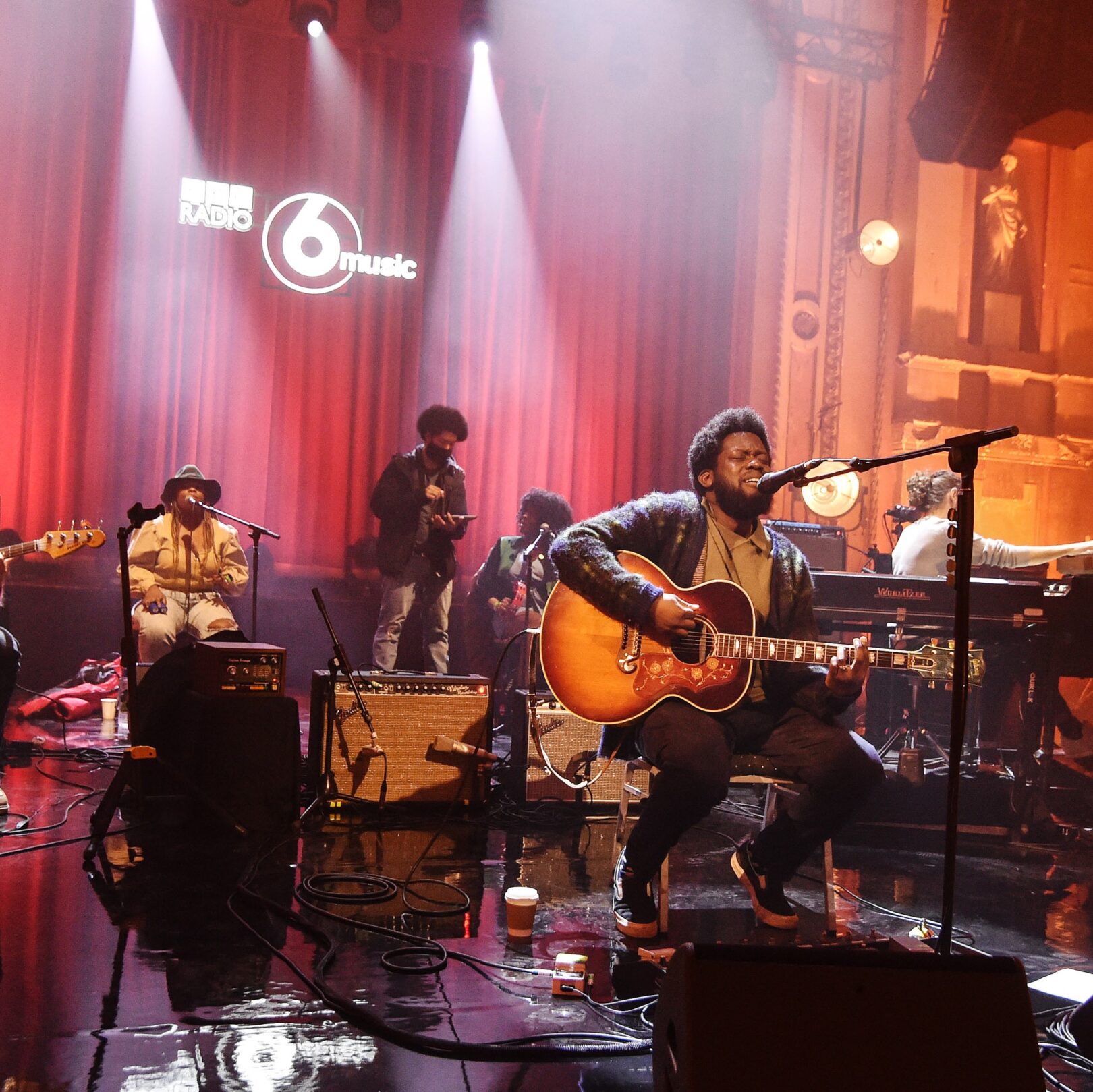 Live band playing music with person playing the acoustic guitar and singing in the foreground whilst people sing and play other instruments behind him, all in front of a red velvet curtain with the BBC 6 music logo behind them.