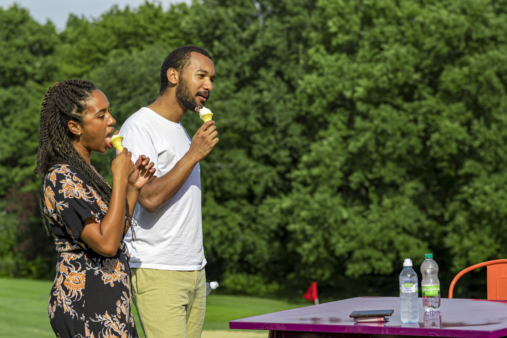 Two people enjoying ice cream cones in the sun by a table on a golf course.