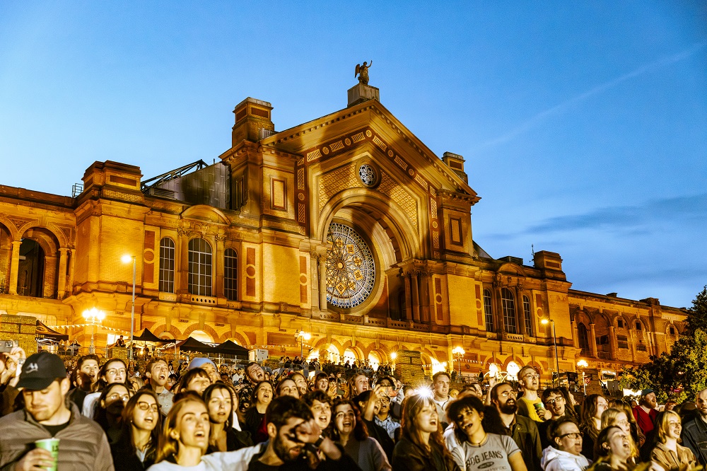 A large group of people in front of a Alexandra Palace singing at sunset.